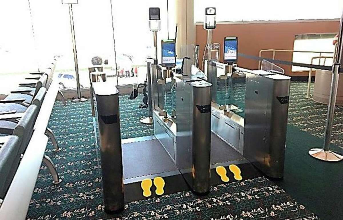 Biometric face recognition boarding gate area with two parallel metal gate units, glass barriers, overhead screens and cameras, yellow footprint floor markers, and waiting area seating in the background