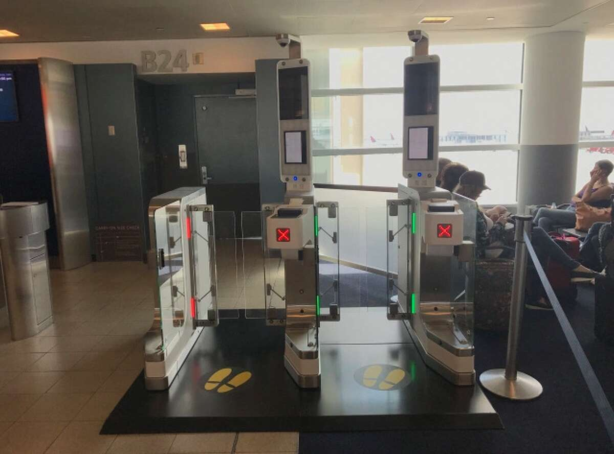 Boarding gate B24 area showing two side-by-side automated gate readers with glass barriers, red X indicators, yellow footprint floor markings, and passengers seated in the lounge to the right by floor-to-ceiling windows