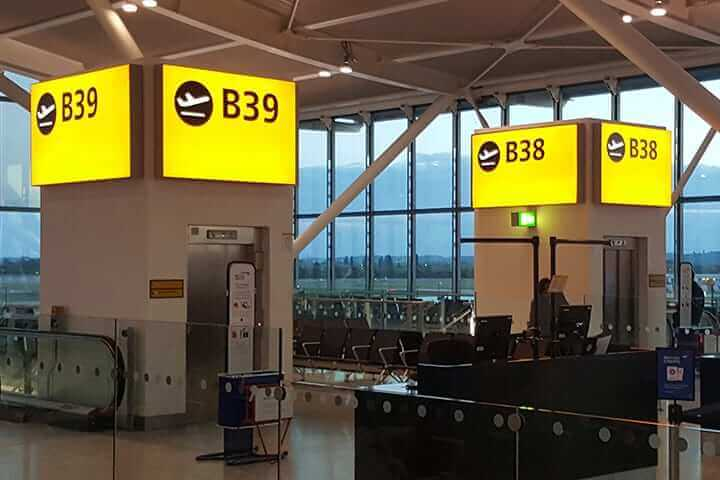 Wide view of a modern airport terminal gate area with bright overhead recessed lighting, gate signage pillars B38 and B39, and large floor-to-ceiling windows showing the airfield beyond