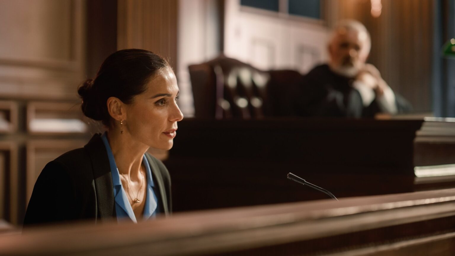 Witness on the stand in a modern American courtroom with warm wooden paneling and overhead judicial lighting