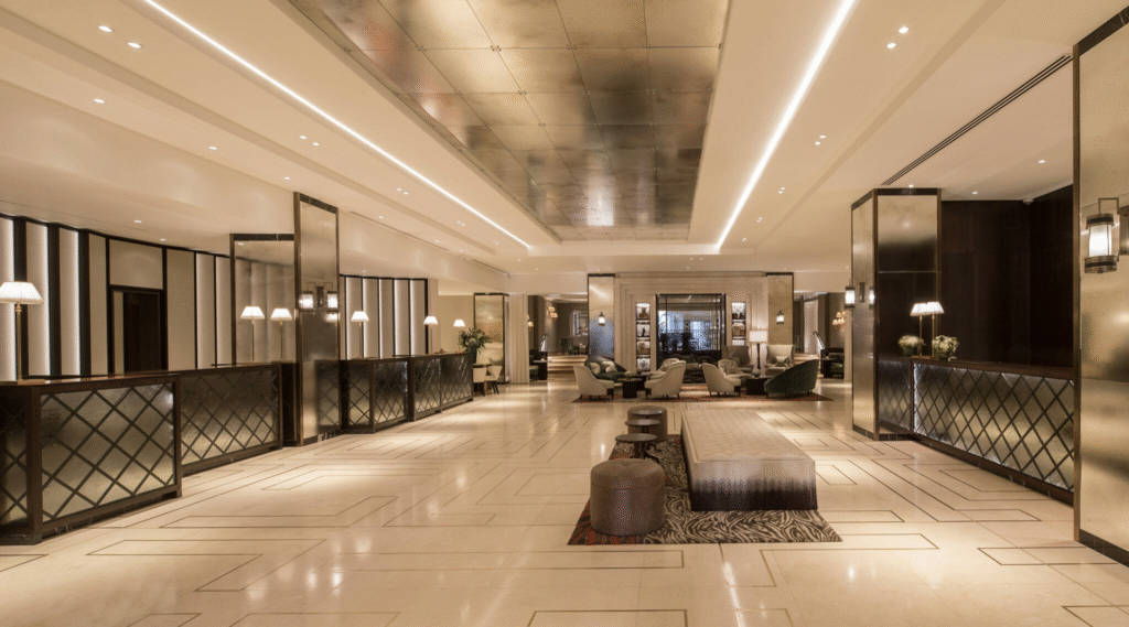 Panoramic view of a luxury hotel lobby with marble flooring, modern chandelier above a central reception desk, geometric patterned side panels, comfortable lounge seating, and abundant warm ambient lighting