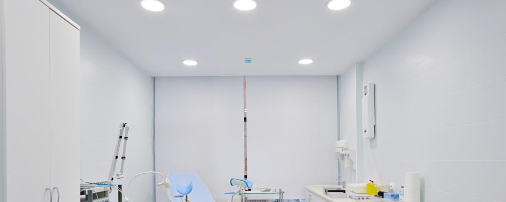 Wide view of a clean modern hospital medical procedure room with a blue patient bed in the center, white walls, ceiling-mounted circular surgical light fixtures providing bright illumination, various medical devices and carts positioned around the room, and a clean light-colored floor in a professional sterile environment