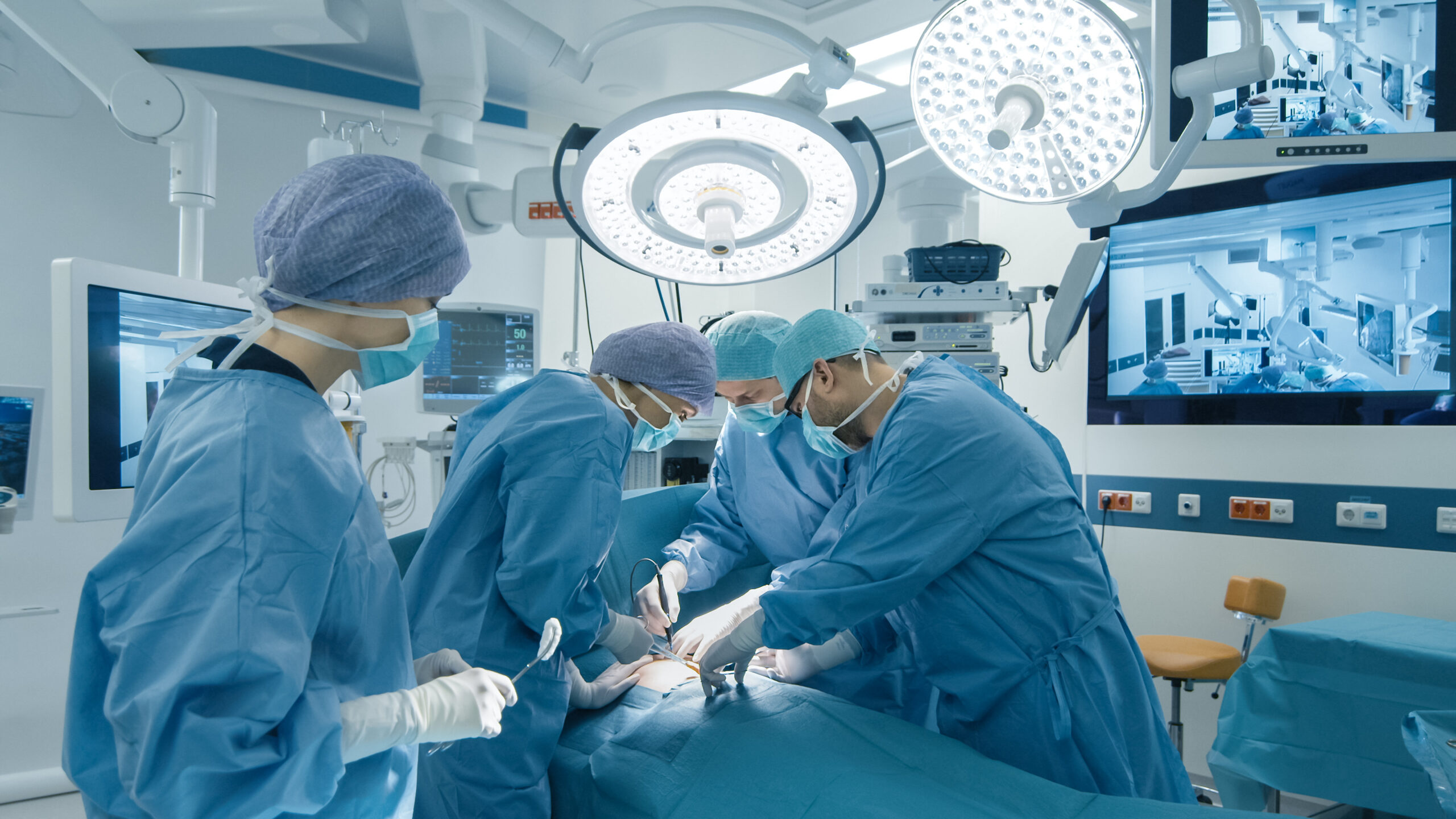 Wide-angle view of a hospital operating room with four surgeons in blue scrubs and caps gathered around a surgical table, two large circular LED surgical light heads hanging from the ceiling providing focused illumination, medical monitors and equipment on the walls, and a clean professional surgical environment