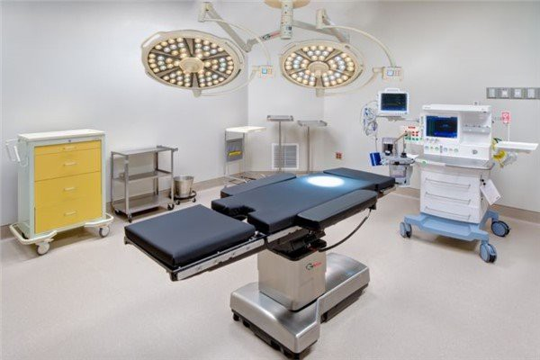 Wide view of a modern hospital operating room with a black surgical table at the center, two large circular LED surgical light heads hanging from the ceiling providing bright focused illumination, a yellow drawer cart on the left, a medical device with a screen on the right, white walls with emergency call buttons, and a beige polished floor in a clean professional environment