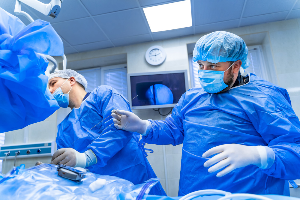 Two surgeons in blue surgical scrubs and caps performing an operation in an operating room, with one surgeon wearing white surgical gloves holding instruments, overhead surgical lights providing focused bright illumination on the surgical field, a clock and medical monitor visible on the back wall, and a window with blinds on the right side