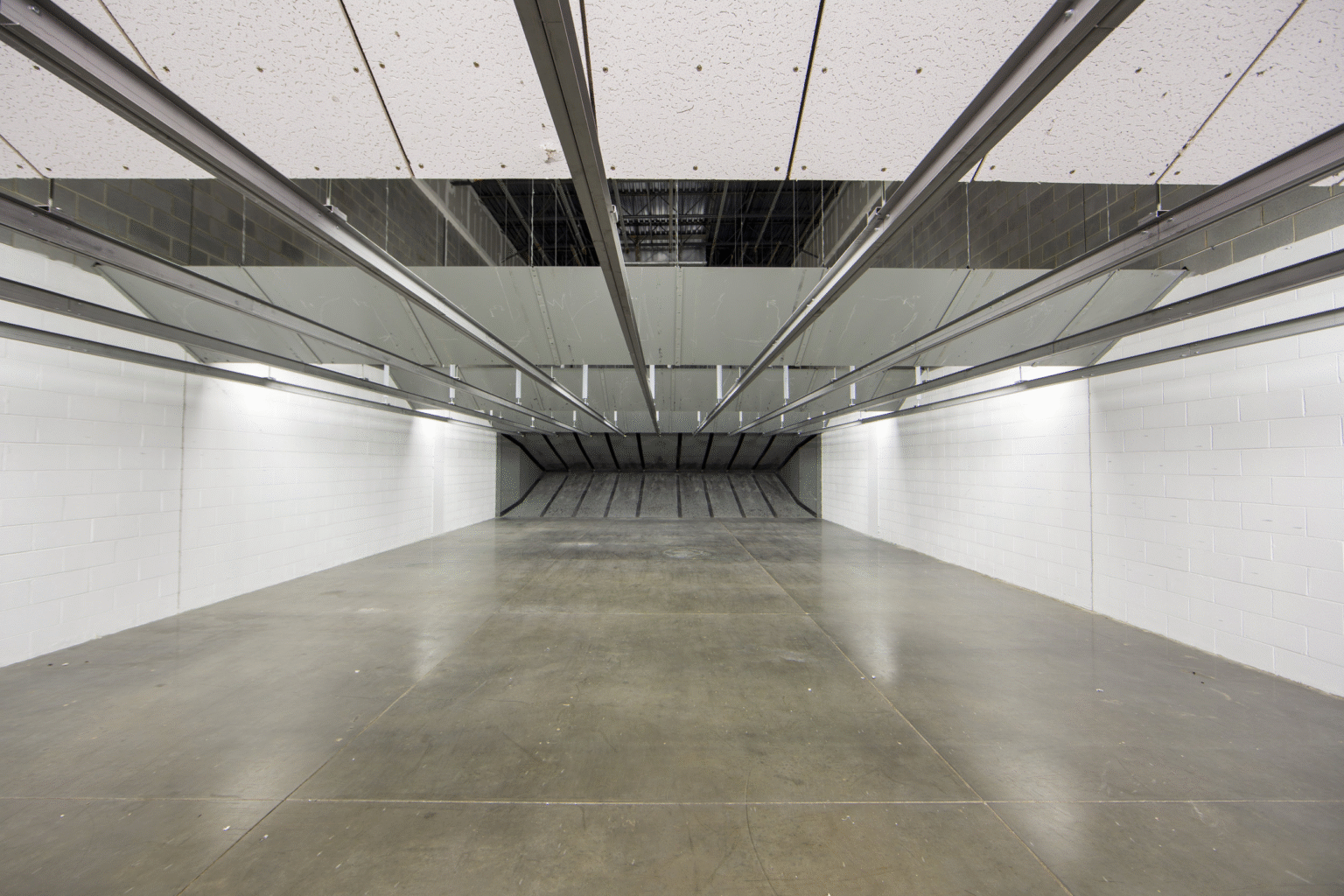 Wide view down the firing lanes of a commercial indoor shooting range with overhead industrial lighting and bullet traps at the far end