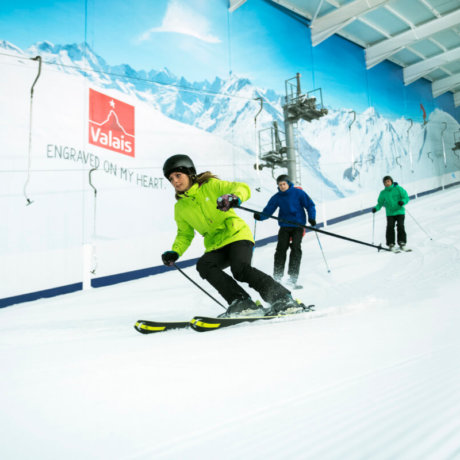 Skier in a green jacket and helmet carving down an indoor snow lesson slope with instructor guidance visible behind