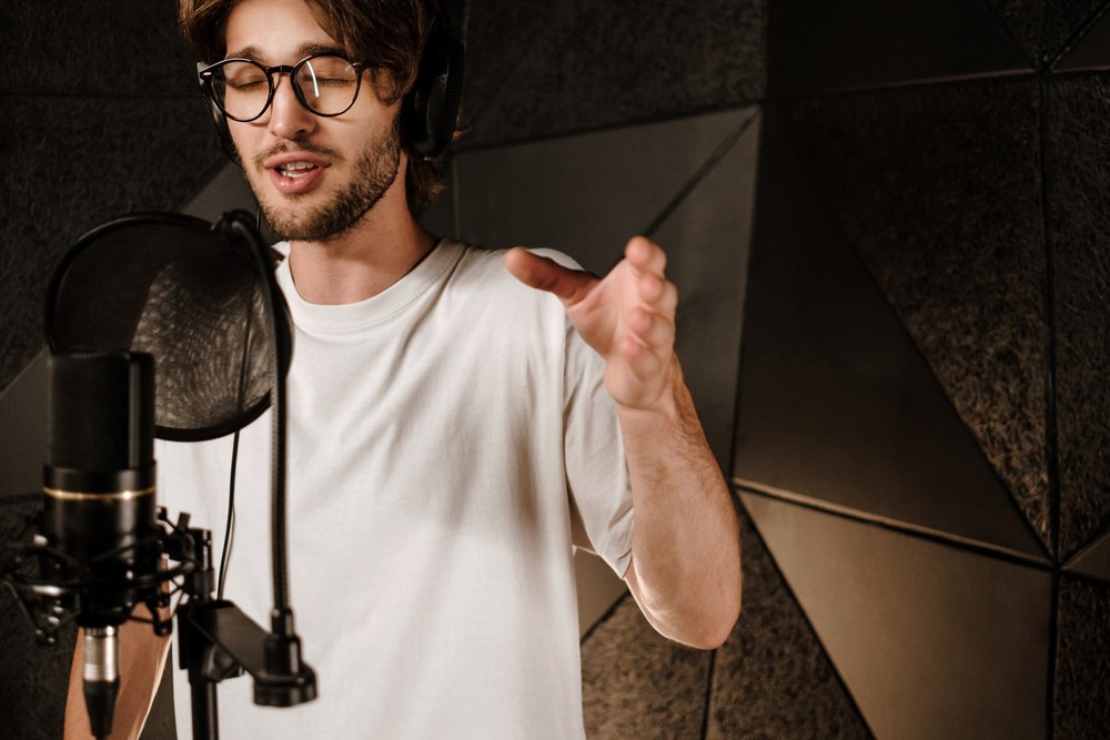 Close-up of a focused musician singing into a studio microphone with warm task lighting and acoustic paneling visible in background